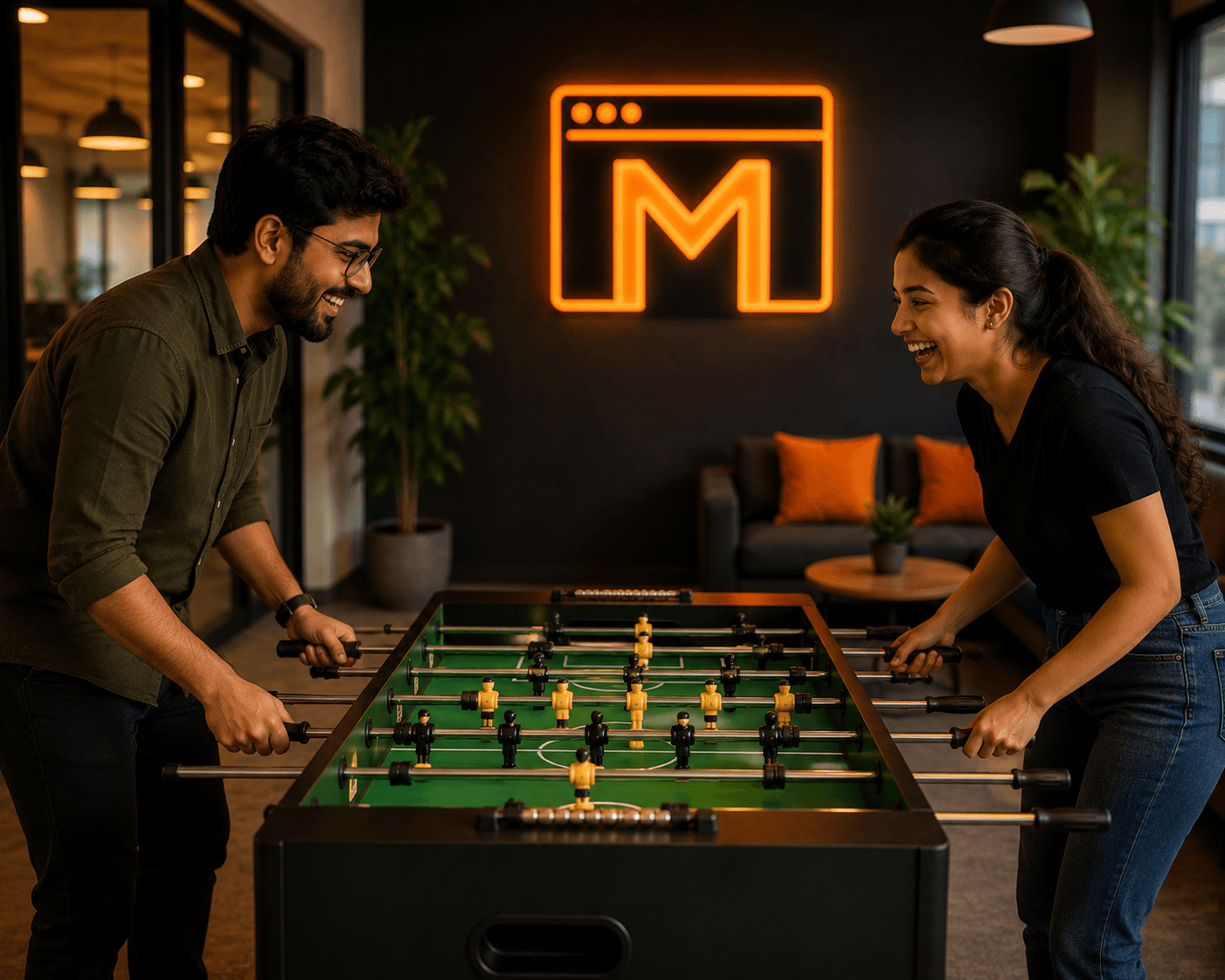 Two Mejona colleagues playing foosball and laughing, with a glowing orange Mejona M neon sign on the back wall of the recreation lounge.